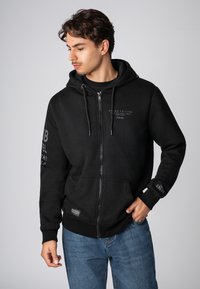 Schwarze Zip-Hoodie aus weichem Stoff mit einer Kapuze mit Kordelzug, vorderen Taschen und bedrucktem Branding auf der Brust und den Ärmeln.