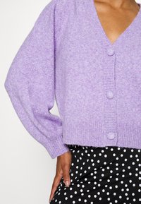 Lila stickad cardigan med V-utsnitt, långa ballongsleeves och fyra runda knappar. Bärs över en svart kjol med prickar.