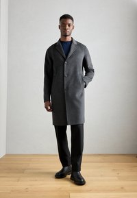 Grijze wollen jas met een notch-kraag, drie knopen en voorzakken, gecombineerd met zwarte broek en zwarte schoenen op een houten vloer.