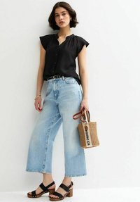 Blusa negra de manga corta con botones, vaqueros de pernera ancha de color azul claro, sandalias negras de tacón cuadrado y un bolso pequeño tejido con una correa.