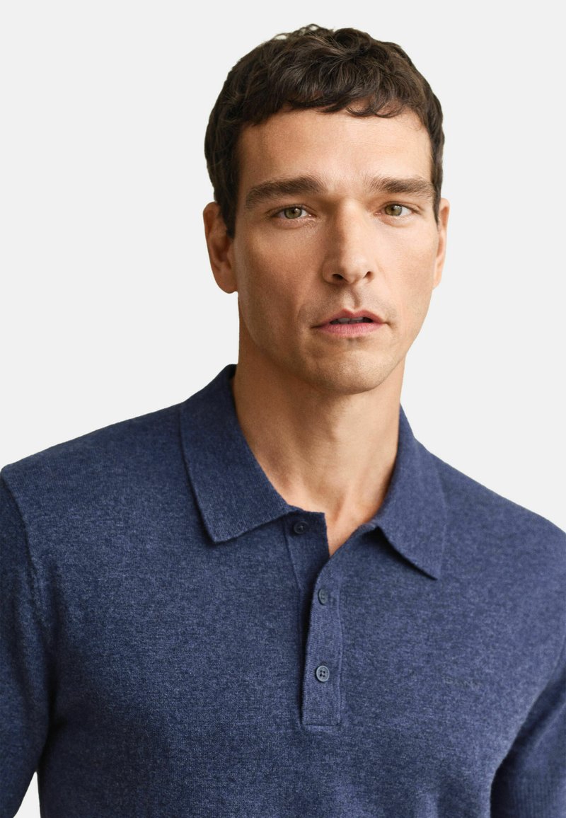 Homme aux cheveux courts et foncés portant un polo bleu marine boutonné, faisant face à l'avant sur un fond clair uni.