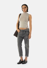 Haut sans manches beige, jeans gris déchirés, chaussures noires avec couture détaillée, et un sac à épaule sombre. Le mannequin se tient devant un fond blanc.