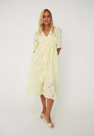 Kvinde iført en lys gul, hulmønstret midi-kjole med puffede korte ærmer og beige åbne hæle med blomsterdetaljer.