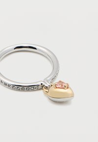 Δαχτυλίδι ασημένιο με μικρές διαυγείς πέτρες τοποθετημένες κατά μήκος της ζώνης και ένα χρυσό μενταγιόν σε σχήμα καρδιάς με ροζ πολύτιμο λίθο επισυνάπτεται.