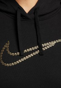 Černá mikina vyrobená z měkké tkaniny, která má zlatě vyšívané logo Nike napříč hrudníkem. Obsahuje šňůrky pro nastavitelnou velikost.