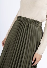Oliven grøn plisseret nederdel med elastisk talje, designet i et blødt, skinnende stof. Stylings med en lys beige langærmet top.