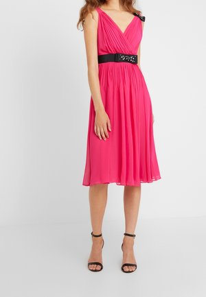 Vrouw draagt een mouwloze, knielange felroze jurk met plooien en een zwarte tailleband met decoratieve studs, gecombineerd met zwarte hakken.