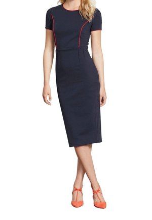 Femme portant une robe bleu marine jusqu'aux genoux avec des détails en biais rouges et des talons hauts orange à lanières, se tenant avec les jambes croisées.