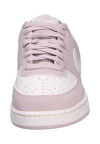 Vista frontal de una zapatilla Nike de color rosa claro y blanco con cordones y una caja de punta perforada, mostrando el logo de Nike en el lado y la lengüeta.
