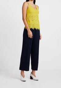 Top sans manches jaune avec des bordures en dentelle, associé à un pantalon large bleu marine et des chaussures à talons carrés blanches, présentant une texture de tissu lisse.