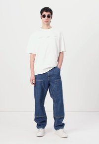Weißes, oversized T-Shirt mit Grafik auf der Vorderseite, kombiniert mit lockeren blauen Jeans. Das Model trägt runde Sonnenbrillen und weiße Sneakers.