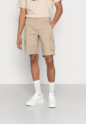 Beige Cargo-Shorts mit Seitentaschen, bis zur Mitte des Oberschenkels. Kombiniert mit weißen Sneakern und knielangen weißen Socken, getragen mit einem hellbeigen T-Shirt.