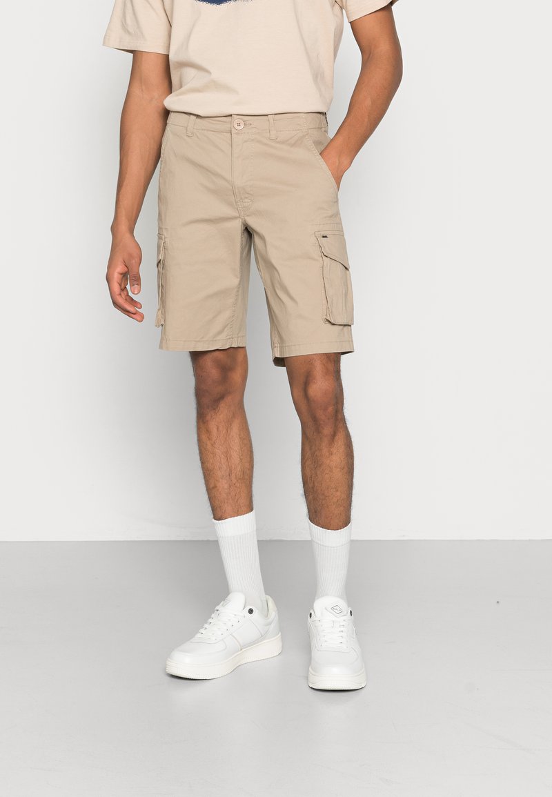 Beige Cargo-Shorts mit Seitentaschen, bis zur Mitte des Oberschenkels. Kombiniert mit weißen Sneakern und knielangen weißen Socken, getragen mit einem hellbeigen T-Shirt.