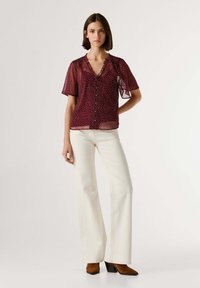 Femme portant un chemisier transparent bordeaux à pois blancs, un pantalon blanc à jambes larges et des bottines marron à talons, debout devant un fond uni.