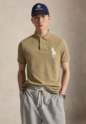 Jeune homme portant un polo beige avec un logo cheval blanc, un pantalon gris à cordon, une casquette bleu marine et une montre-bracelet, debout, mains dans les poches.