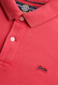 Polo rojo con textura, con dos botones blancos abrochados y un pequeño logo bordado de "SuperDry" en el pecho.