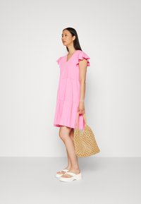 Vestido rosa com decote em V, mangas com babados e design em camadas, combinado com uma bolsa de palha e sandálias brancas de plataforma. Tecido de textura suave.