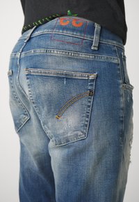 Jeans i denim med en bleknad blå tvätt, med en röd och orange logotyp på bakfickan, samt kontrasterande gyllene sömmar på en av fickorna.
