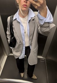 Femme dans un ascenseur portant un blazer gris, une chemise bleu clair, un top blanc, un pantalon noir, des chaussures à motif serpent et un sac à main noir, prenant un selfie.