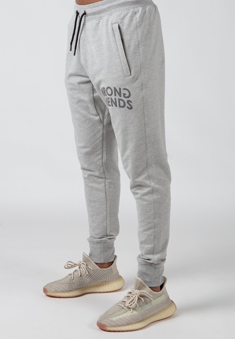 Grå sweatpants med sidelommer og elastiske manchetter, med "WRONG FRIENDS" trykt i sort på det øverste lår. Båret med beige sneakers.