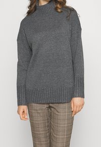Eine Frau trägt einen dunkelgrauen gerippten Rollkragenpullover und beige karierte Hosen, während sie vor einem schlichten hellen Hintergrund steht.