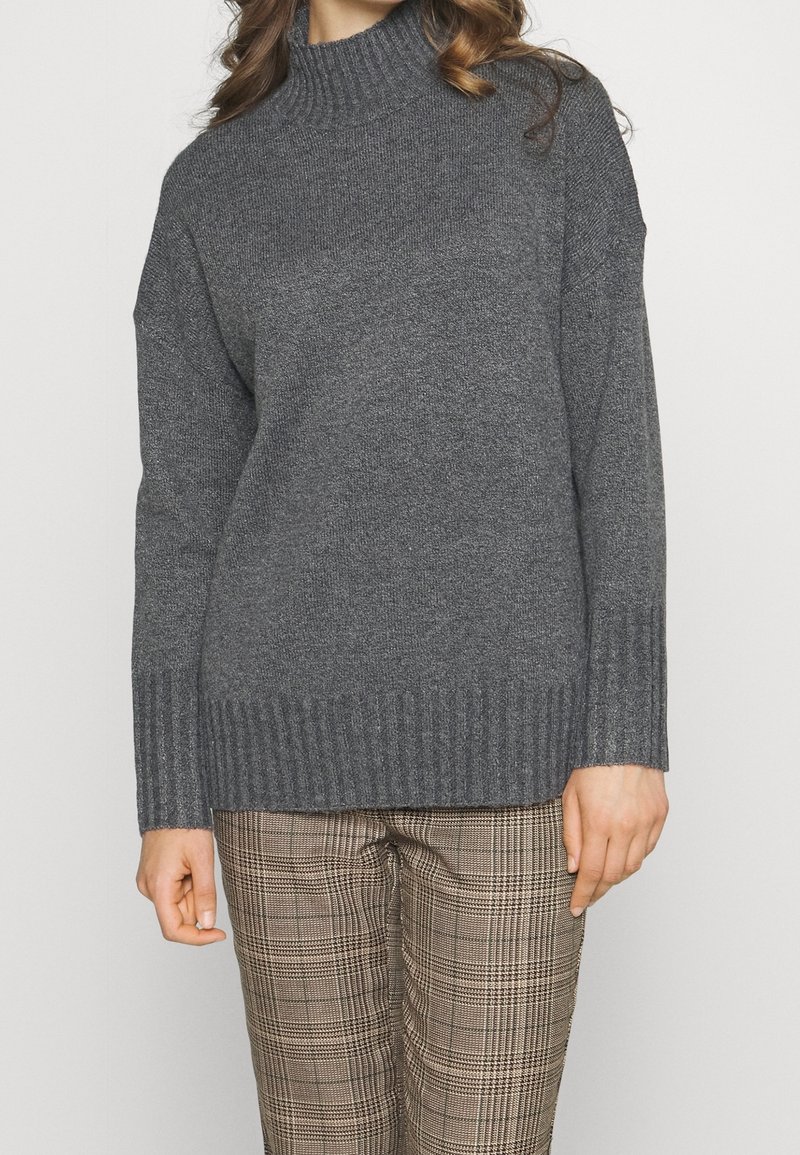 Eine Frau trägt einen dunkelgrauen gerippten Rollkragenpullover und beige karierte Hosen, während sie vor einem schlichten hellen Hintergrund steht.