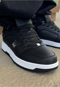 Svart lær sneaker med perforert tåboks, hvit mellomsåle og svarte snørebånd. Har en krone-logo på siden og teksturerte detaljer.