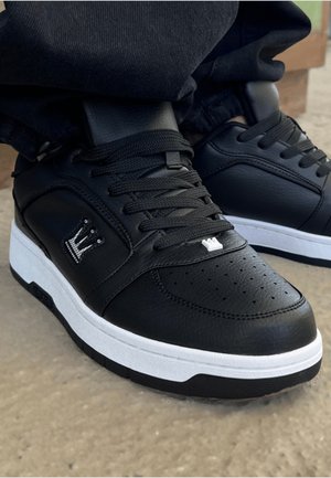 Svart lær sneaker med perforert tåboks, hvit mellomsåle og svarte snørebånd. Har en krone-logo på siden og teksturerte detaljer.