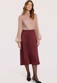 Kobieta ubrana w przejrzystą bluzkę w odcieniu różowego, z długimi rękawami, wsuniętą w spódnicę midi w burgundowym kolorze o wysokim stanie, z czarnymi przejrzystymi rajstopami i spiczastymi szpilkami.