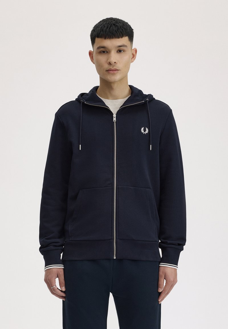 Jonge man draagt een marineblauwe hoodie met ritssluiting en een klein wit lauwerkranslogo op de borst, staand tegen een effen achtergrond.