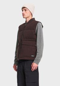 Jeune homme portant un bonnet beige en maille, un gilet matelassé marron avec des poches, un pull gris à manches longues et un pantalon cargo noir, regardant vers la gauche sur fond blanc.