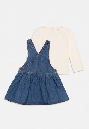 Chemise crème à manches longues associée à une robe salopette en denim bleu avec des bretelles croisées dans le dos et des boutons sur fond blanc.