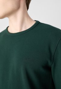 Tamnozeleni džemper od vaflaste pletenine s okruglim izrezom. Ima suptilni logo "BOSS" u istoj boji, naglašavajući teksturiranu tkaninu i detalje šivanja.