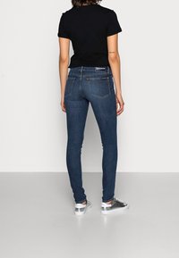 Vaqueros skinny de denim en azul oscuro con bolsillos traseros, costuras doradas y una etiqueta negra. Combinados con zapatillas plateadas y una camiseta negra ajustada.