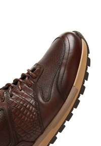 Brown leather sneaker with textured accents, perforated sections, and a tan rubber sole. Features brown laces and a padded collar.