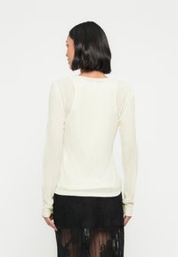 Γυναίκα με κοντά μαύρα μαλλιά φορεί ένα διάφανο κρεμ μακρυμάνικο τοπ και μαύρη δαντελένια φούστα, γυρισμένη με την πλάτη προς ένα απλό λευκό φόντο.