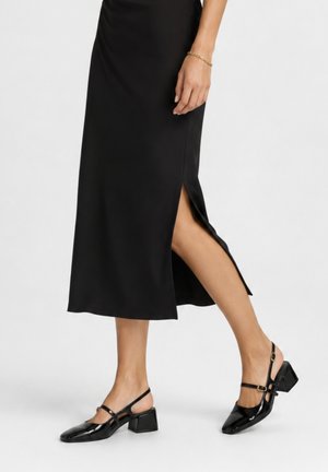 Vrouw draagt een zwarte midi-rok met een zijsplit en zwarte lakleren Mary Jane schoenen met lage hak terwijl ze loopt op een witte achtergrond.