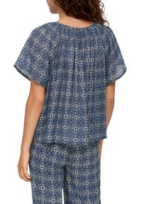 Haut bleu à motifs avec manches courtes, encolure smockée et coupe ample, présentant des motifs floraux sur un fond géométrique.