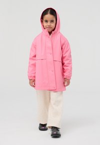 Giacca impermeabile rosa con cappuccio, bottoni a pressione, polsini elastici, abbinata a pantaloni color crema con un motivo sottile e scarpe nere.