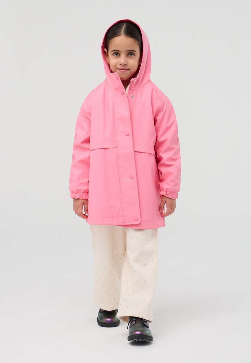 Giacca impermeabile rosa con cappuccio, bottoni a pressione, polsini elastici, abbinata a pantaloni color crema con un motivo sottile e scarpe nere.