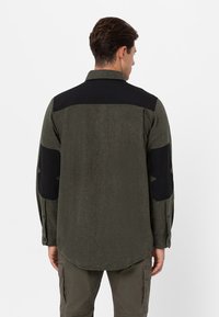 Camicia in flanella verde con accenti neri sulle spalle e sui gomiti, maniche lunghe e orlo curvo. Tessuto testurizzato con un motivo sottile.