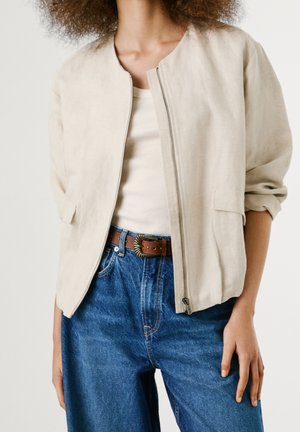 Femme portant une veste beige zippée par-dessus un haut blanc, associée à un jean bleu taille haute et une ceinture marron avec une boucle décorative.