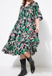 Femme portant une robe noire à fleurs avec des feuilles roses et vertes, associée à des bottes de cowboy noires mi-mollet brodées, sur fond blanc.