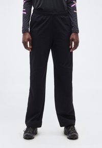 Άτομο που φοράει μαύρο παντελόνι χαλαρής εφαρμογής, μαύρα παπούτσια τρεξίματος σε μονοπάτια και μαύρο μακρυμάνικο τοπ με ροζ και κίτρινα σχέδια στα μανίκια.