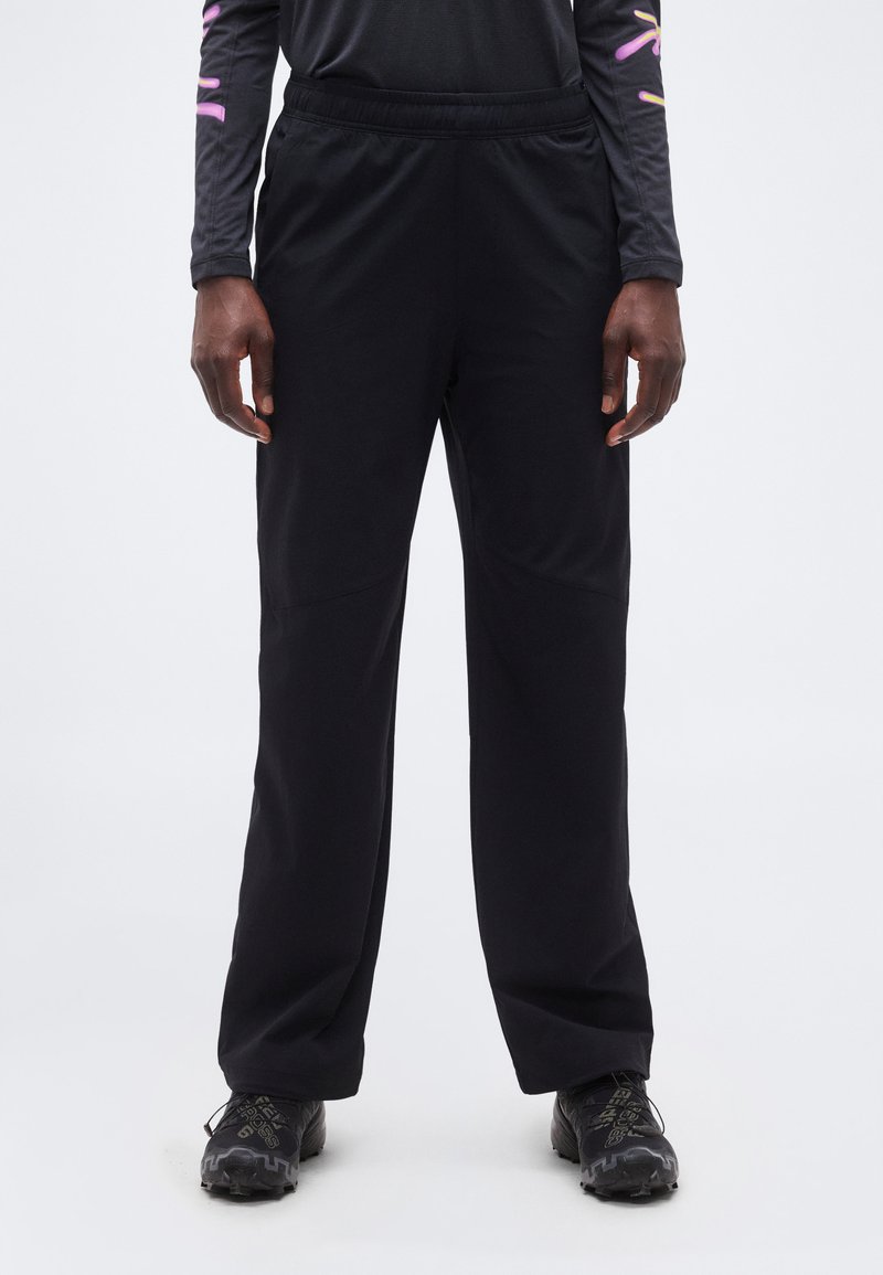 Άτομο που φοράει μαύρο παντελόνι χαλαρής εφαρμογής, μαύρα παπούτσια τρεξίματος σε μονοπάτια και μαύρο μακρυμάνικο τοπ με ροζ και κίτρινα σχέδια στα μανίκια.