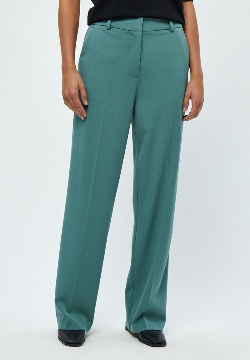 Personne portant un pantalon large taille haute bleu sarcelle et des chaussures noires, debout les mains détendues sur les côtés devant un fond uni.