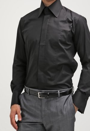 Mann trägt ein eng anliegendes schwarzes Hemd, das in graue Anzugshosen mit schwarzem Ledergürtel gesteckt ist, Hände entspannt seitlich.