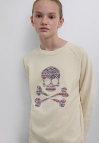 Sweatshirt couleur crème avec un motif de tête de mort et os croisés en motifs géométriques multicolores. Tissu doux avec manches raglan.