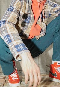 Camicia a quadri nei toni beige, blu e arancione indossata sopra un hoodie arancione, abbinata a pantaloni teal e sneakers arancioni con accenti grigi.