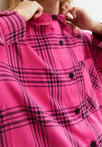 Chemise à carreaux rose en tissu doux, avec des lignes noires, un col et des boutons noirs sur le devant. Détail de poche poitrine notable.